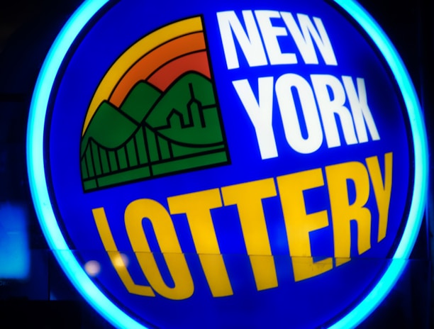 A bright, circular neon sign with a blue background, featuring the words 'New York Lottery' in bold white and yellow letters. The sign includes a colorful illustration of a landscape with green mountains, an orange-red sunset, and a bridge, all encapsulated within the circle.