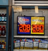 An advertisement display in a store window features lottery jackpot amounts for Mega Millions and Powerball, both showing jackpots of 208 million and 200 million respectively. Below the display, several lottery tickets and promotional posters are visible. To the left of the display, there is a refrigerator stocked with various drinks including different flavors of sports drinks and bottled water.