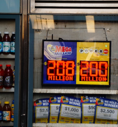 An advertisement display in a store window features lottery jackpot amounts for Mega Millions and Powerball, both showing jackpots of 208 million and 200 million respectively. Below the display, several lottery tickets and promotional posters are visible. To the left of the display, there is a refrigerator stocked with various drinks including different flavors of sports drinks and bottled water.