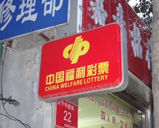 A red sign displaying the text 'China Welfare Lottery' in both Chinese and English, mounted on the exterior of a building. The surroundings include a blue sign with Chinese characters and a partial tree trunk visible on the right side.