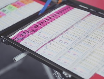 A collection of lottery tickets is organized neatly in a metal tray. The tickets display various numbers and have color-coded sections, primarily in pink and white. Some tickets have vibrant green sections, adding contrast to the overall layout.