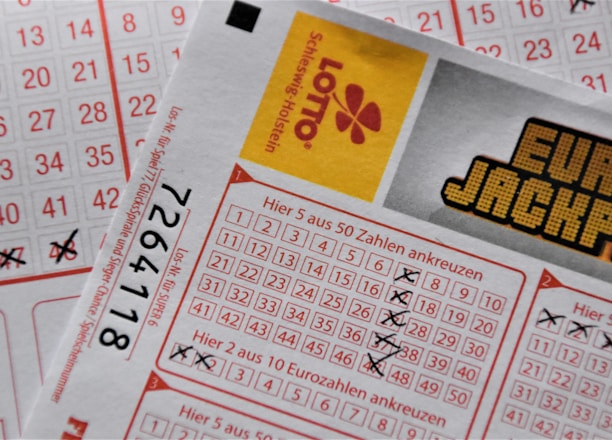 A close-up image of lottery tickets featuring crossed-out numbers, brand logos, and various numbers in a grid format. The tickets have sections for different games, and numbers are marked with black crosses.