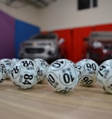 A collection of white lottery balls with black numerals scattered on a wooden surface. Two vehicles are blurred in the background, parked in a room with multicolored walls including teal, purple, blue, and red panels.