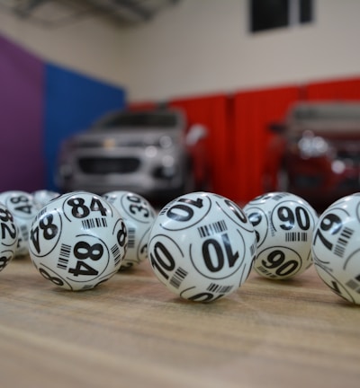 A collection of white lottery balls with black numerals scattered on a wooden surface. Two vehicles are blurred in the background, parked in a room with multicolored walls including teal, purple, blue, and red panels.