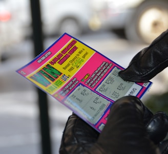 A person wearing black gloves is holding a colorful lottery scratch-off ticket. The ticket includes information about a $1,000,000 guaranteed minimum payout and features multiple games with various prize amounts.