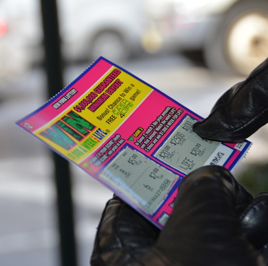 A person wearing black gloves is holding a colorful lottery scratch-off ticket. The ticket includes information about a $1,000,000 guaranteed minimum payout and features multiple games with various prize amounts.