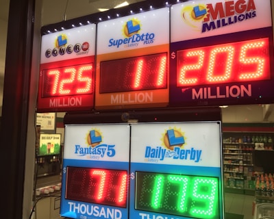 A signboard inside a convenience store displays the current jackpot amounts for various lottery games. There are bright, illuminated numbers in red and green showing the prize amounts: Powerball is at 725 million, Super Lotto Plus at 11 million, Mega Millions at 205 million, Fantasy 5 at 71 thousand, and Daily Derby at 179. In the background, shelves with various drinks and products are visible, along with promotional posters.